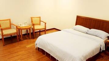 Habitación de hotel con una cama de madera, dos sillas, una mesita con una bandeja y una pared blanca.