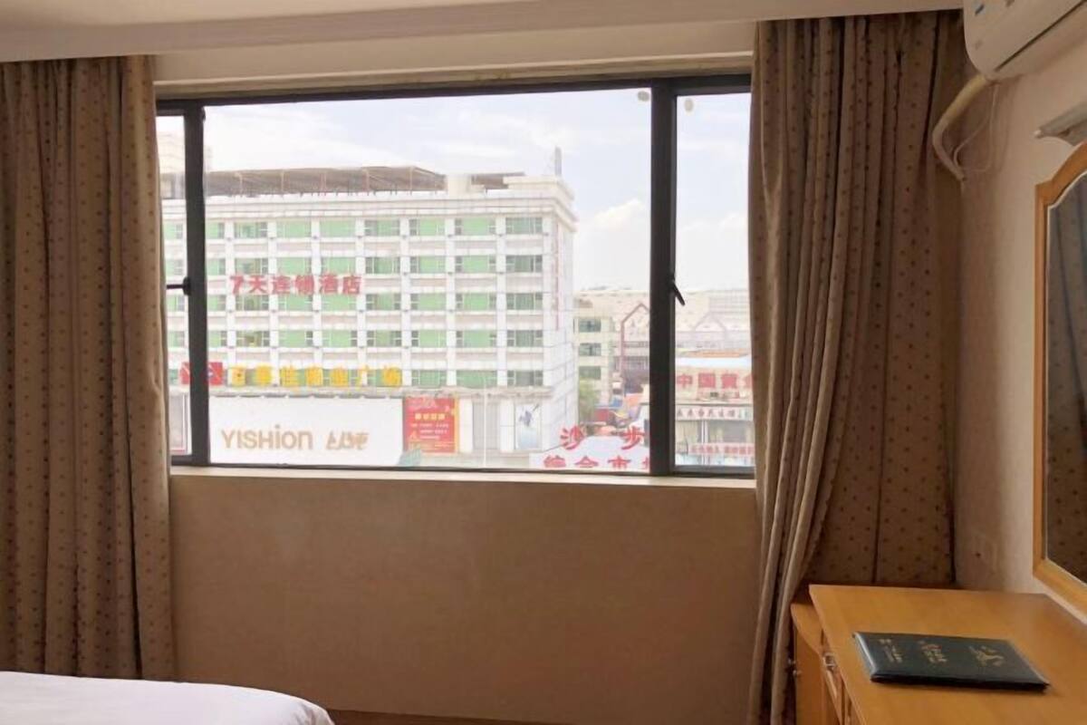 Ein Hotelzimmer mit großem Fenster, Vorhängen, einem Bett, einem Holfnachtkästchen und einer Wand-Klimaanlage.