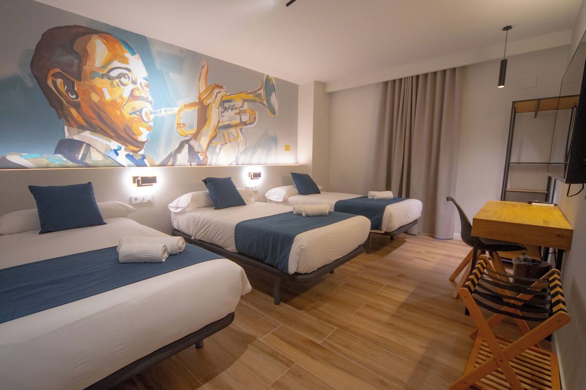 A hotel room with three beds, a wooden floor, a painting on the wall, and a small table with a chair.