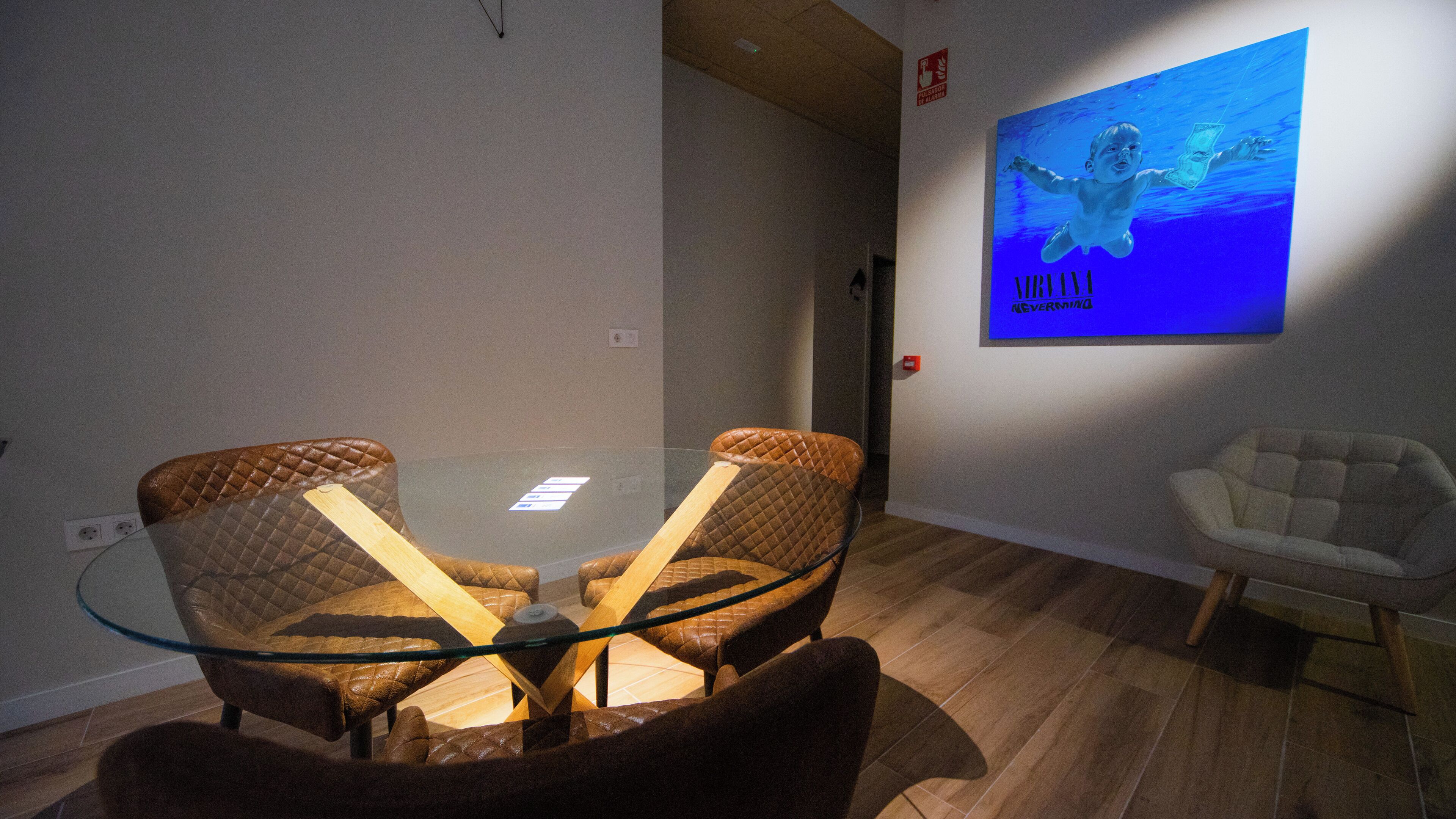 A room with a glass-top table surrounded by two brown leather chairs, a white chair, and a blue painting on the wall.