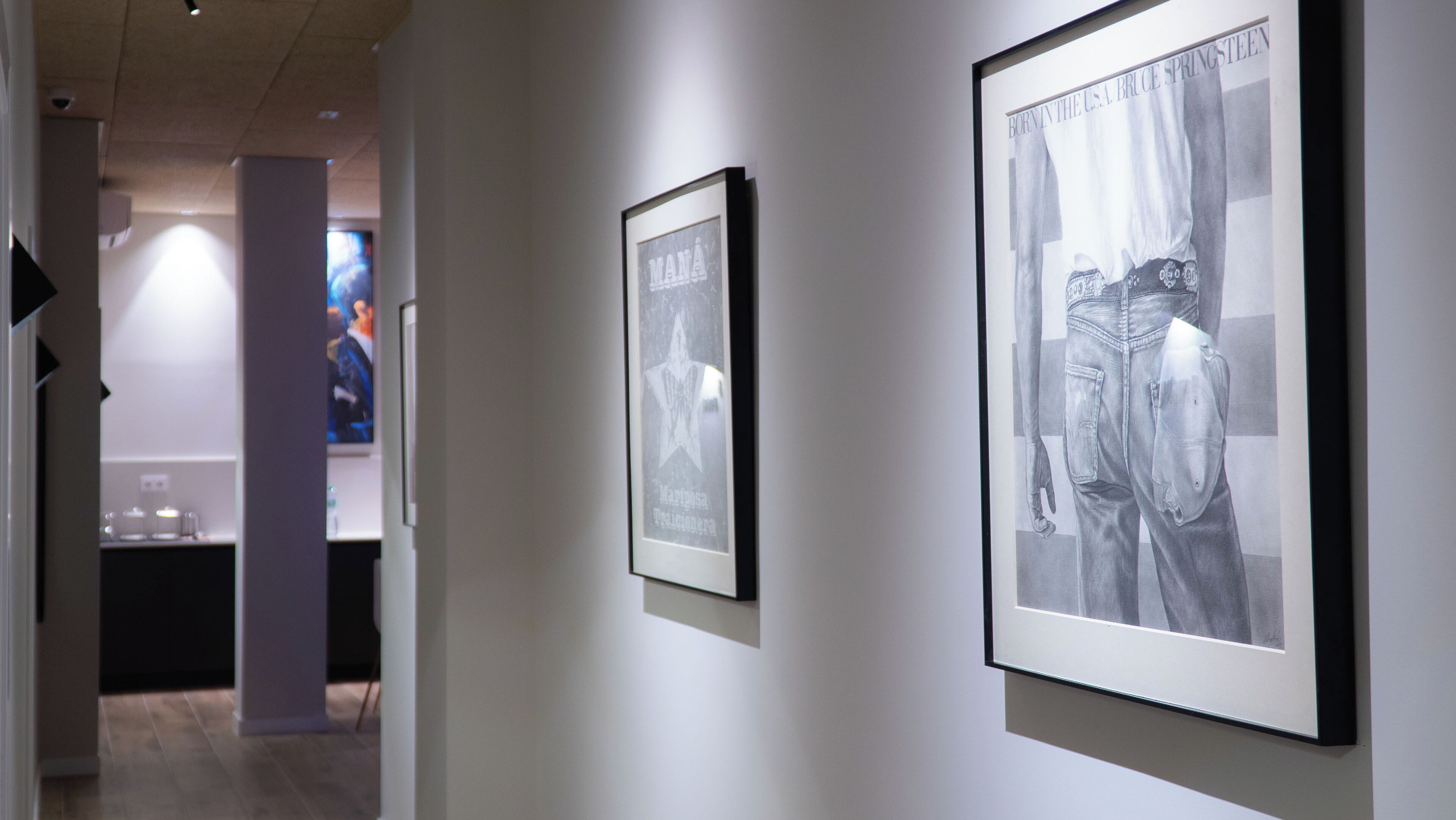 A hallway with framed artwork on the walls, a television screen in the background, and a door leading to another room.