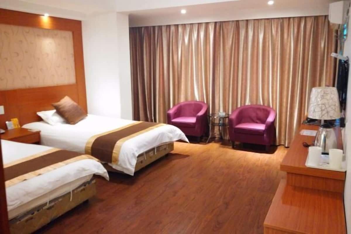 A hotel room with two beds, a wooden headboard, a desk with a lamp, and two pink armchairs.