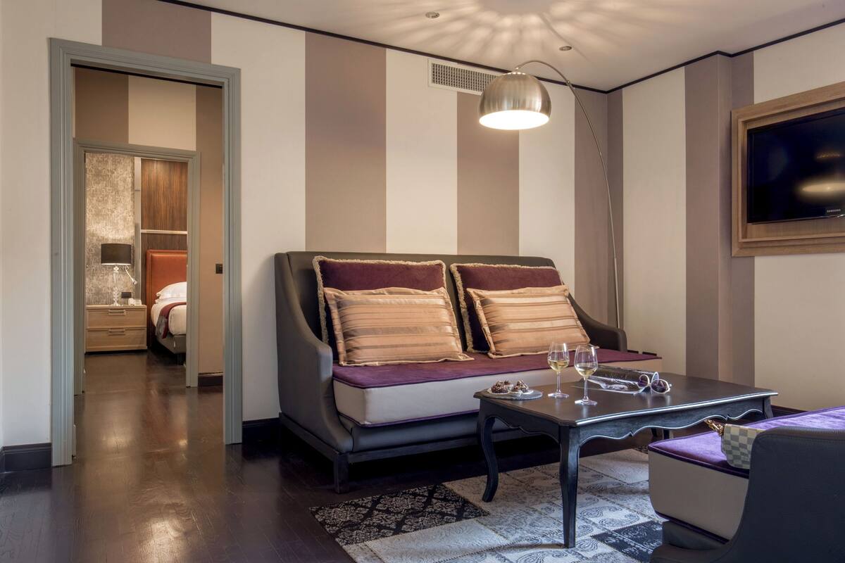 Una habitación de hotel moderna con un sofá, una pequeña mesa con una copa de vino y un televisor en la pared.