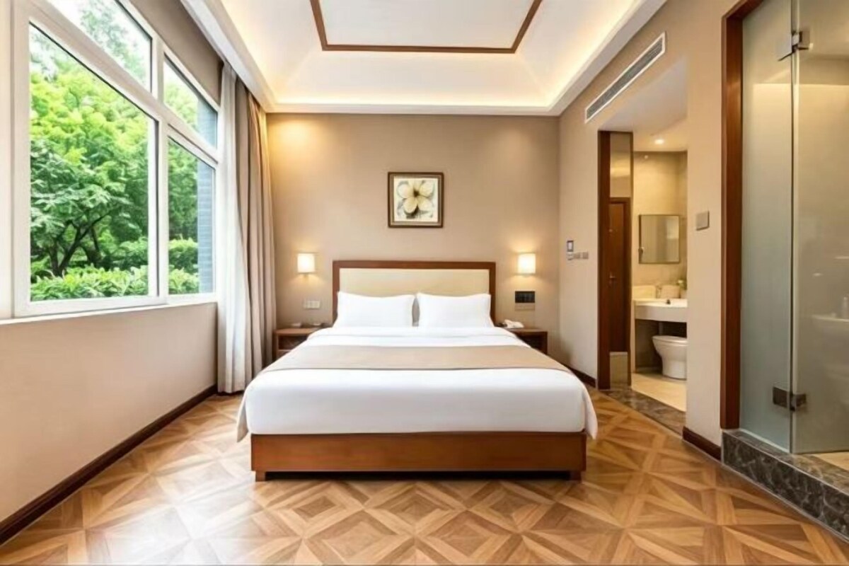 A hotel room with a large bed, bedside lamps, a window with a view of greenery, a shower area, and a wooden floor.