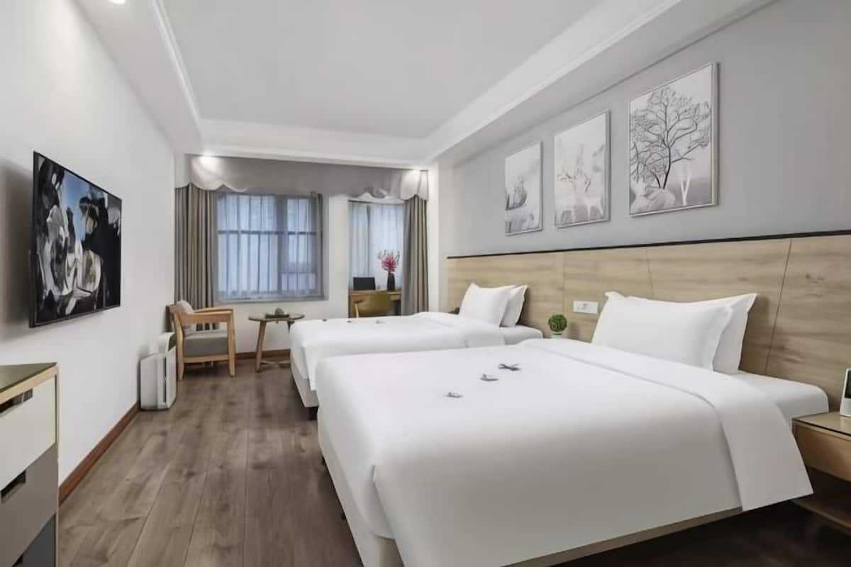A hotel room with two beds, a desk, a chair, a window with curtains, and artwork on the walls.