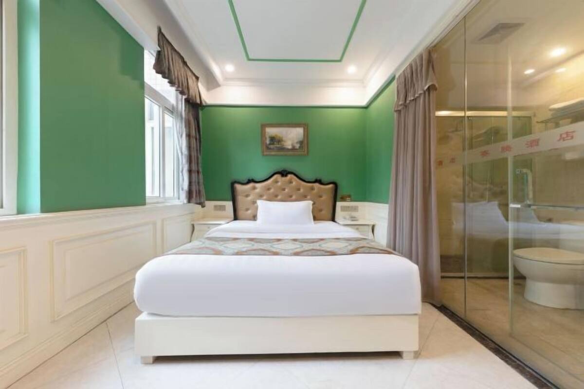 A hotel room with a bed, a nightstand, a window with curtains, and a view of a bathroom through a glass partition.