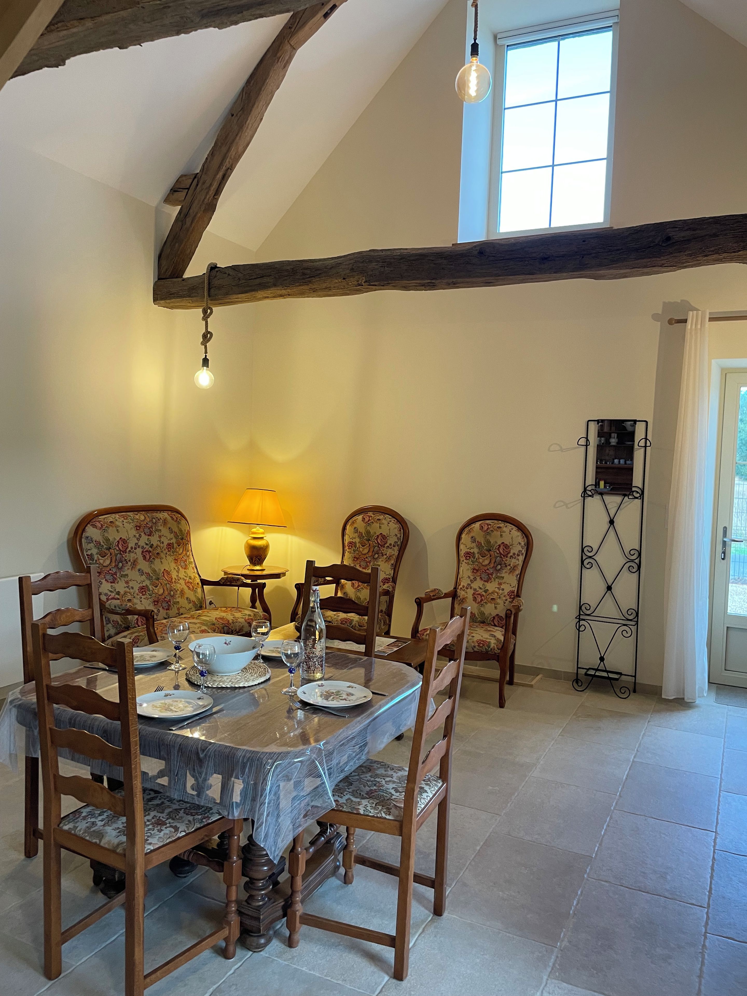 Uma área de refeições com mesa de madeira posta para quatro pessoas, cercada por cadeiras estofadas com motivos florais. Uma luminária oferece iluminação suave, e um suporte de metal preto e alongado é visível na parede.
