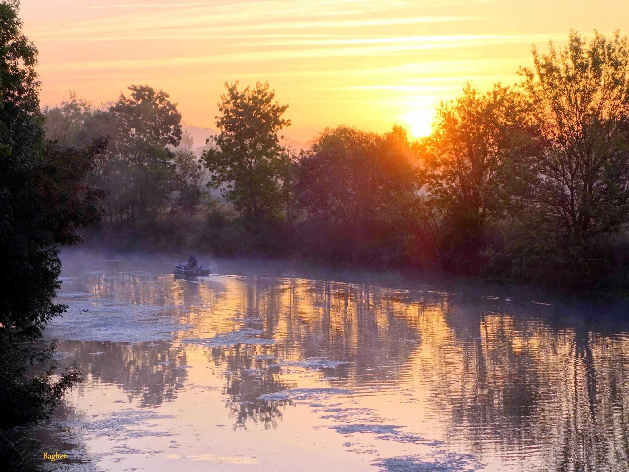 A serene river scene at sunset with a boat, surrounded by trees and reflecting the sky's colors.