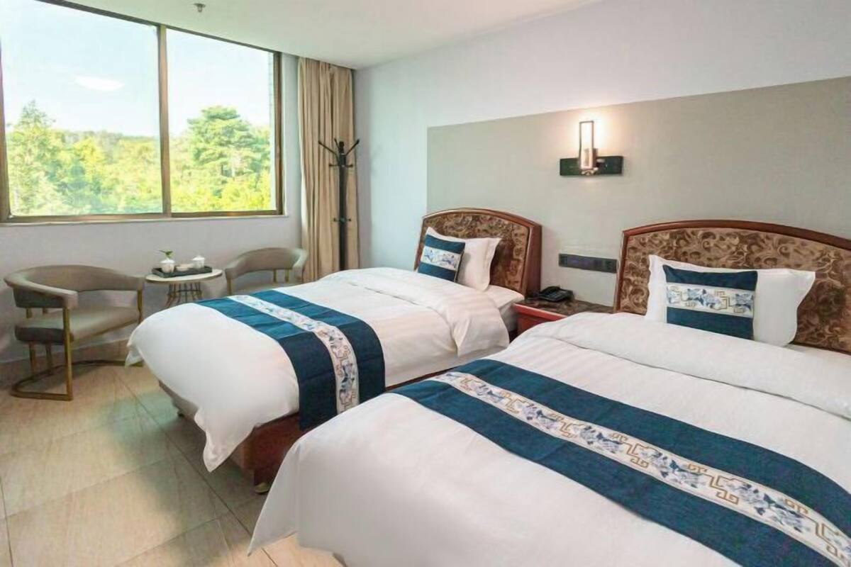 A hotel room with two beds, a small table, a chair, and a large window with a view of trees.