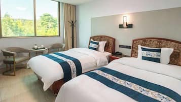 A hotel room with two beds, a small table, a chair, and a large window with a view of trees.