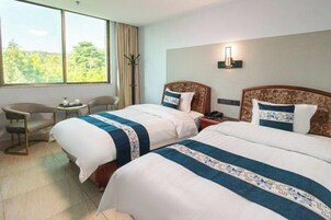 A hotel room with two beds, a small table, a chair, and a large window with a view of trees.