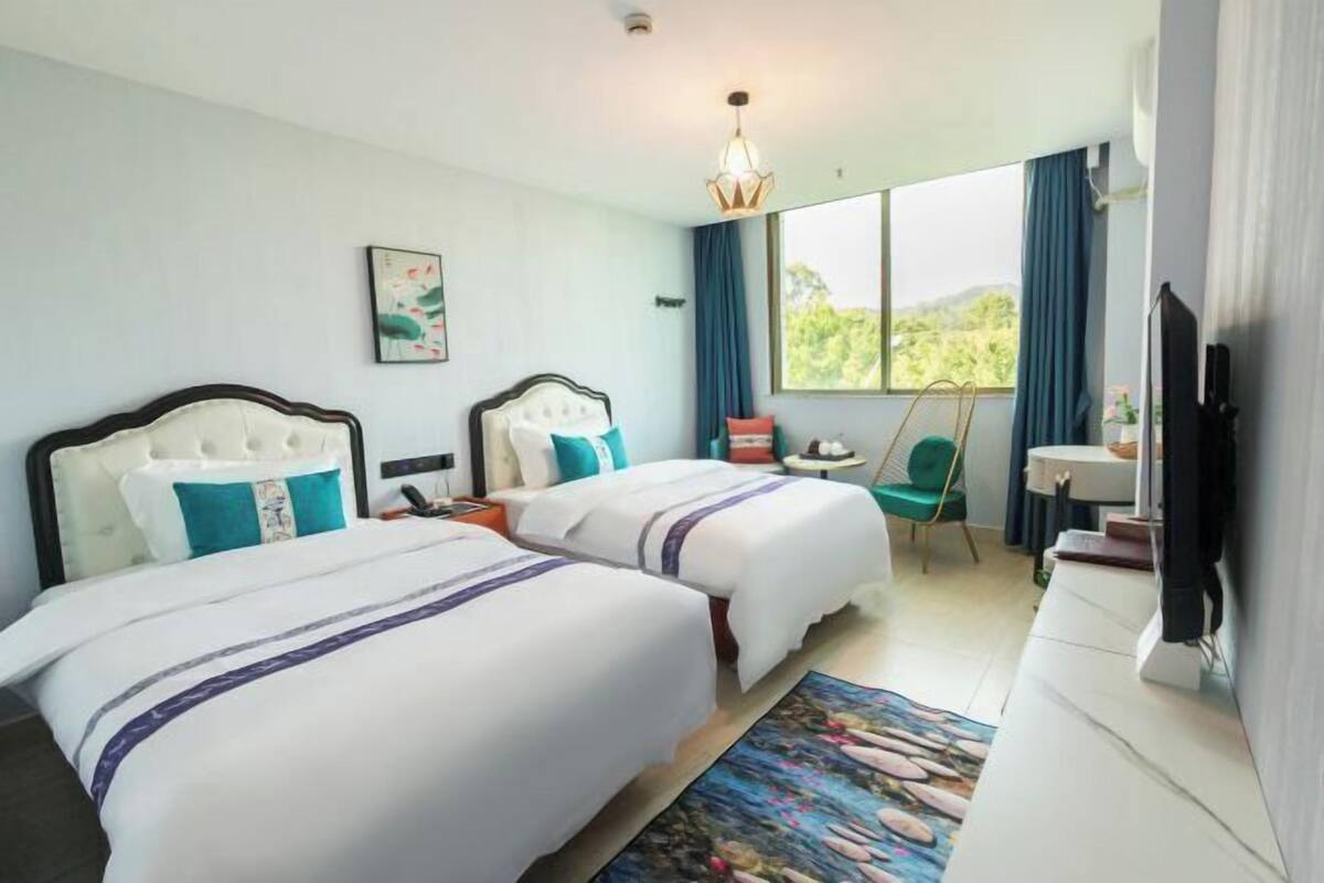 A hotel room with two beds, a desk, a chair, a television, and a window with a view of greenery.
