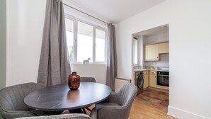 A modern kitchen with a washing machine, a dining area with a round table and grey chairs, and a window with grey curtains. - The Hackney Apartment (London)