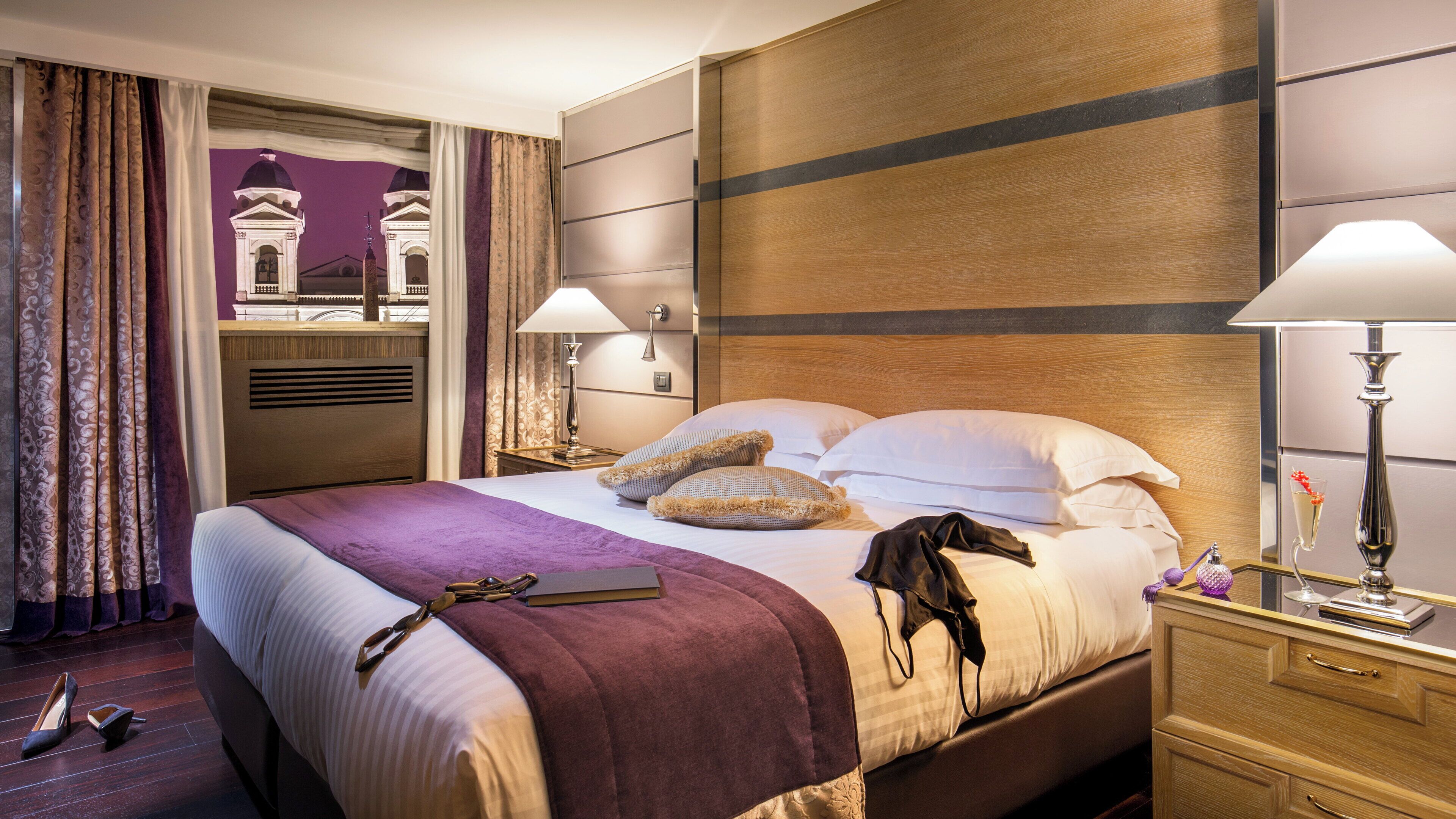 A hotel room with a large bed, two bedside tables, a nightstand, a lamp, and a view of a building through the window.