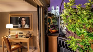 A hotel room with a desk, chair, lamp, and a television. There is a balcony with plants and a view of a building with a clock tower. - The Inn at the Spanish Steps Exclusive Collection - The Nest at The Spanish Steps (Rome)
