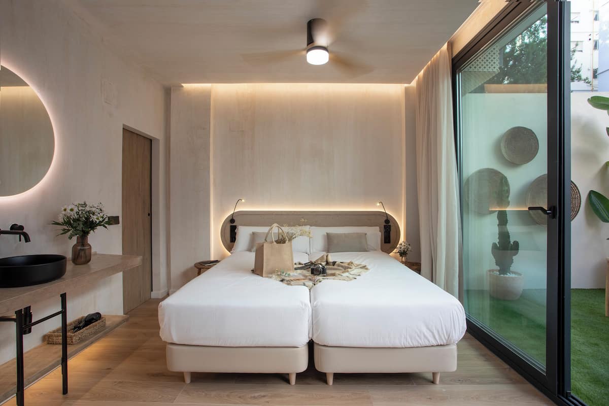 A modern bedroom with a large bed, bedside tables, a round mirror, and a glass door leading to a balcony with plants.