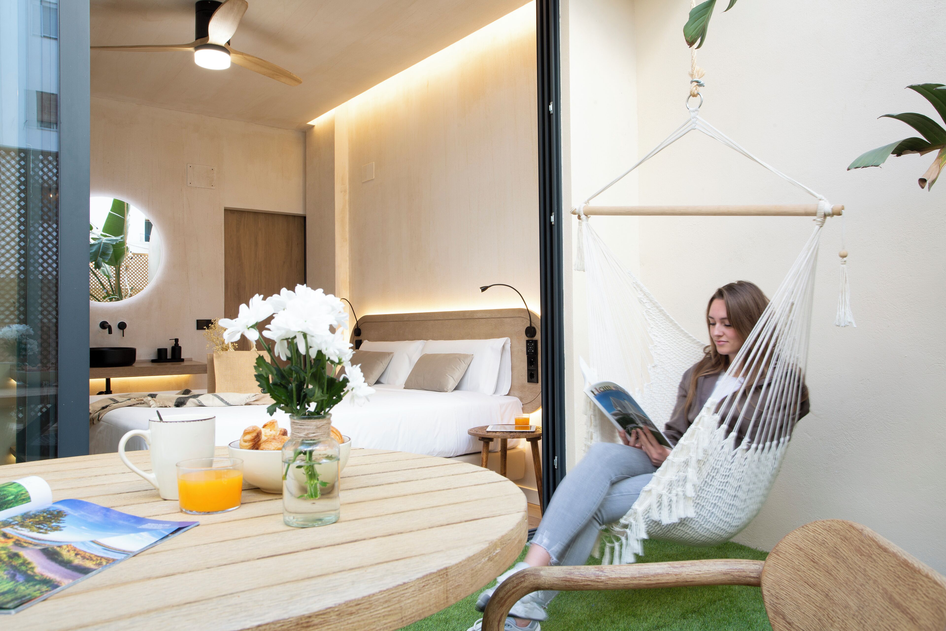 Une chambre d’hôtel moderne avec un lit, une table ronde en bois sur laquelle sont disposés des aliments pour le petit-déjeuner, une chaise blanche et un hamac.