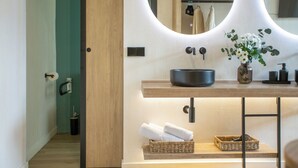 Un baño con tocador de madera, espejo, lavamanos y un florero con flores.