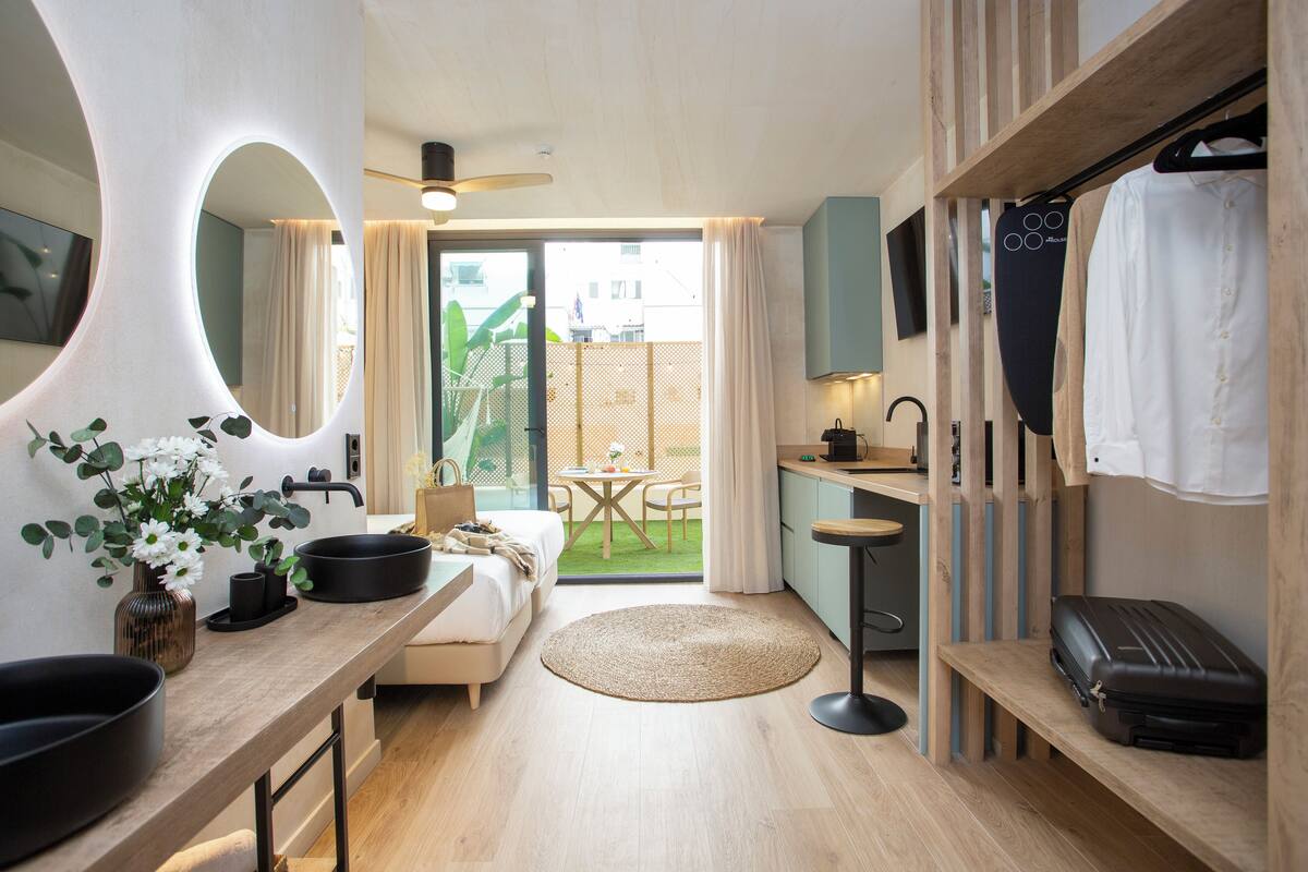 A modern hotel room with a bed, a round dining table, a kitchen area, and a balcony with a view of the outside.