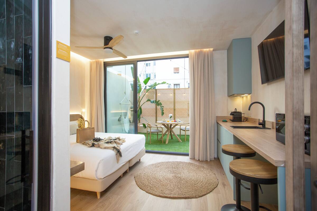 A modern hotel room with a bed, a small kitchen area, a dining table, and a view of the outdoors.