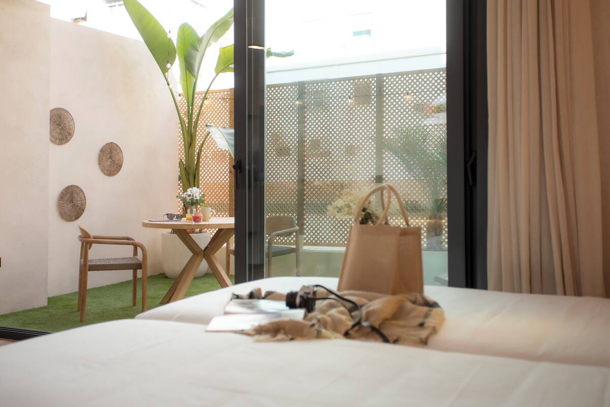 A hotel room with a bed, a small table with a chair, a potted plant, and a view through a sliding door.