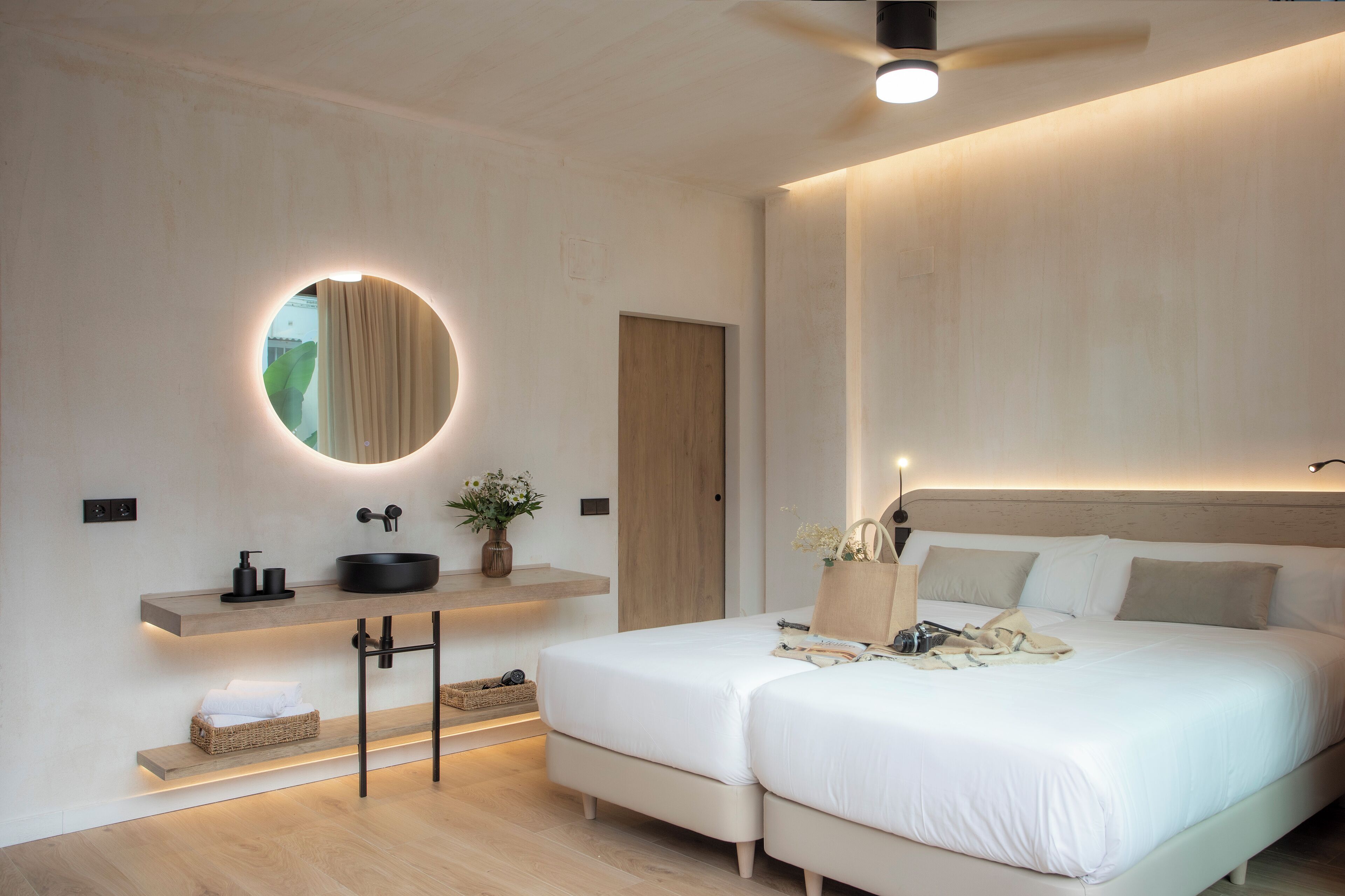 Une chambre moderne avec un lit double, des tables de chevet, un miroir rond et une étagère intégrée où sont rangés les articles de toilette.