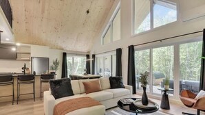 Ein geräumiger Wohnbereich mit hoher Decke, großen Fenstern, einer Couch, einem Couchtisch und einer Küchenzeile.