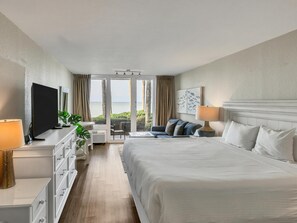 A hotel room with a bed, a TV, a desk with a lamp, a chair, a sofa, and a view of the sea. - Bayfront Walkout Studio with Sunset Views at Sandestin (Miramar Beach)