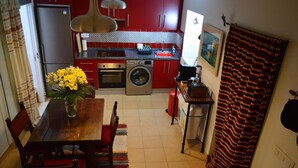 Une cuisine compacte avec des meubles rouges, un réfrigérateur en acier inoxydable, un lave-linge et un coin repas comprenant une table et des chaises en bois.