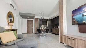 A modern living room with a grey sofa, a dining table with chairs, a kitchen area with dark cabinets, and a mounted TV displaying a scenic view.