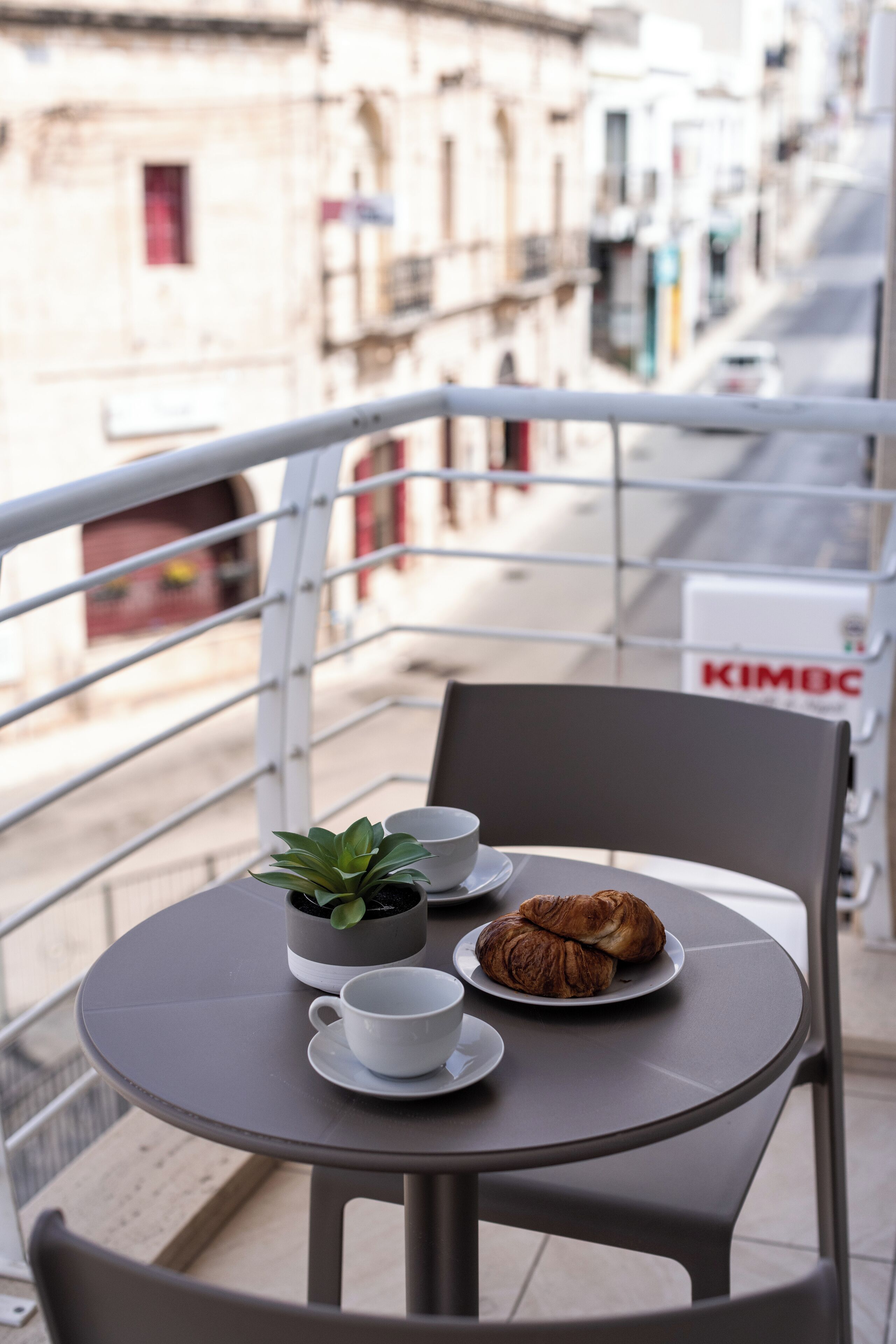 Un balcón con una mesa dispuesta para dos personas, incluyendo una planta, tazas y croissants.
