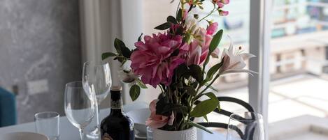 Una mesa de comedor preparada con platos, tazones y copas de vino blancos, una botella de vino y un florero con flores rosas.