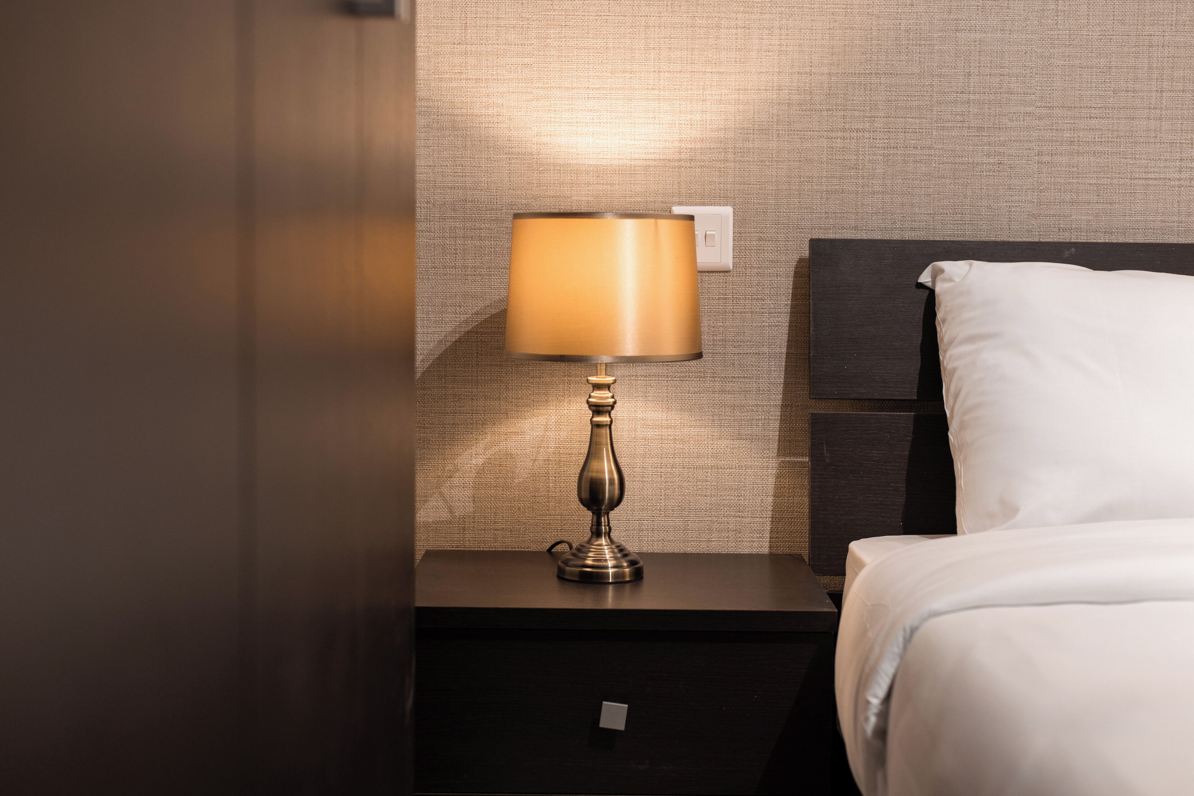 A neatly made bed with white sheets, a dark wooden nightstand with a lamp, and a textured wall behind.