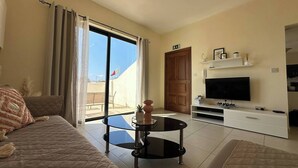Ein modernes Wohnzimmer mit einer Couch, einem Glastisch, einem Flachbildfernseher und einer Schiebetür zu einem Außenbereich.