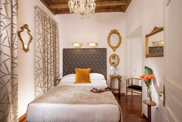 A hotel room with a large bed, a chandelier, a mirror, and a small table with a vase of flowers.