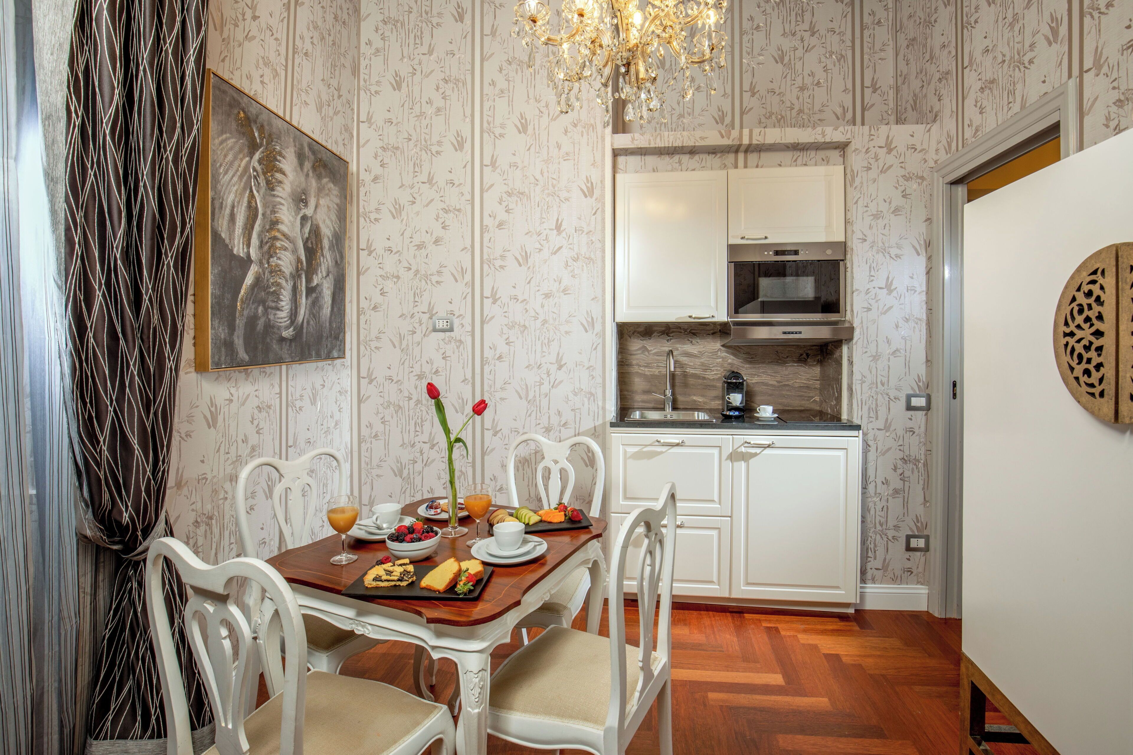 Zona pranzo con tavolo in legno, sedie bianche e un quadro raffigurante un elefante. Sullo sfondo, una cucina con mobili bianchi e una cappa in acciaio inossidabile.