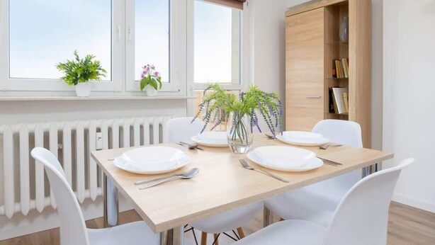 A dining area with a table set for four, white chairs, a radiator, a window with blinds, and a wooden cabinet with books. - Air-conditioned Apartment | Close to the Royal Baths (Warszawa)
