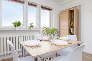 Ein Essbereich mit einem Tisch, gedeckt für vier Personen, weißen Stühlen, einem Heizkörper, einem Fenster mit Jalousien und einem Holzschrank mit Büchern.