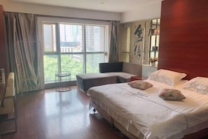 A hotel room with a large bed, a small seating area, and a view of greenery through the window.
