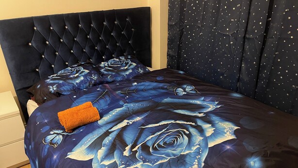 A bed with a dark headboard, a floral bedspread, and a folded orange towel. - 4 Beds, Sleeps 15, Near Lcy Airport & Excel London (London)