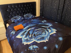 A bed with a dark headboard, a floral bedspread, and a folded orange towel. - 4 Beds, Sleeps 15, Near Lcy Airport & Excel London (London)