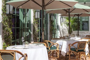 Un espacio de comedor al aire libre con mesas dispuestas para dos, sillas de mimbre y grandes sombrillas que dan sombra.