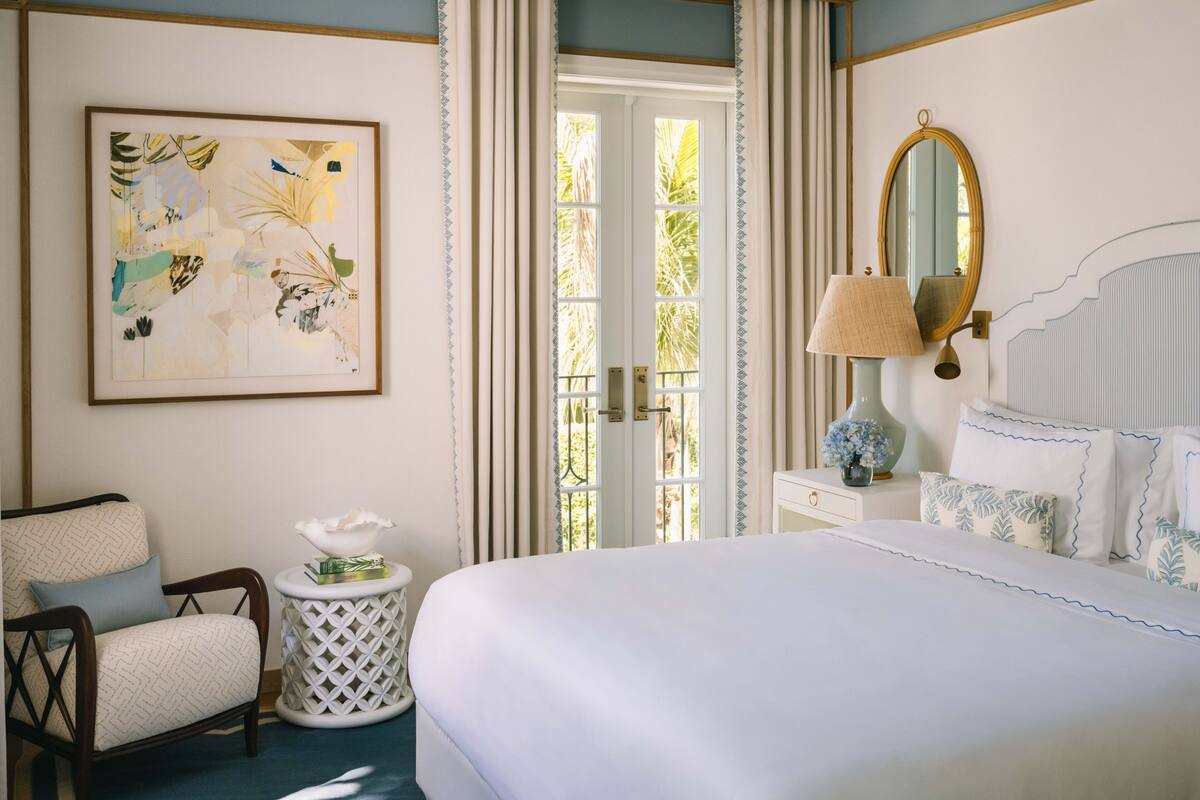 A hotel room with a bed, a chair, a small side table, a lamp, a mirror, and a door leading to a balcony.