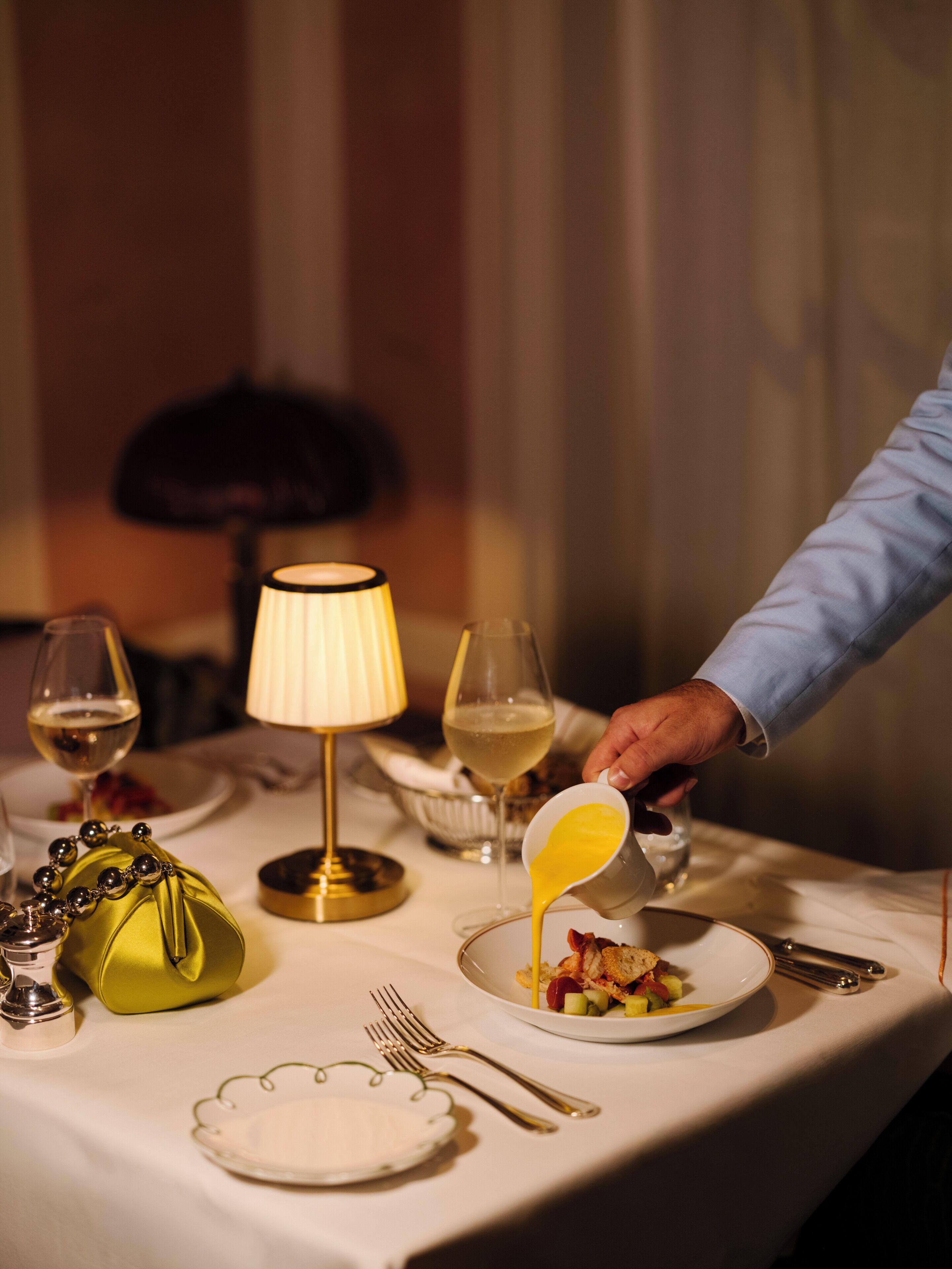 Una mesa de comedor con una lámpara, copas de vino, un cuenco de fruta y una persona sirviendo una bebida.