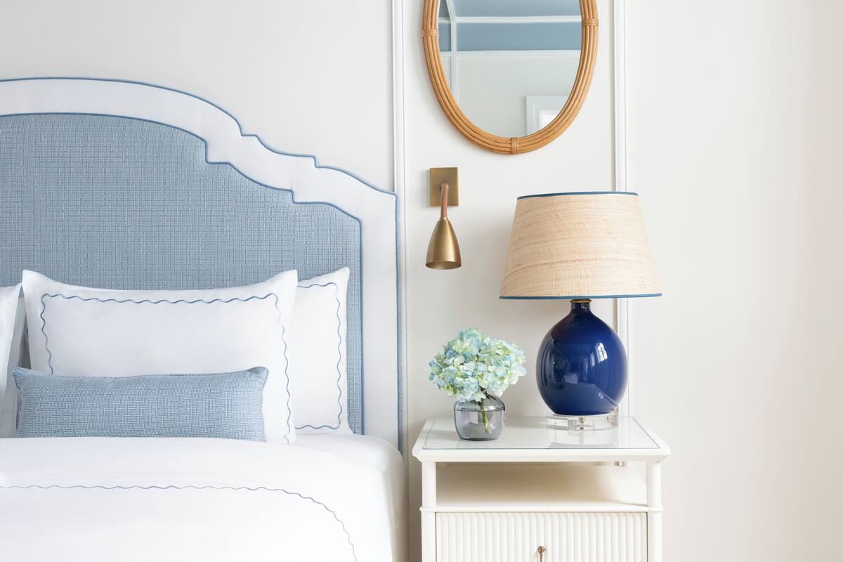 A bed with a headboard, a bedside lamp, a mirror, and a small table with a vase of flowers.