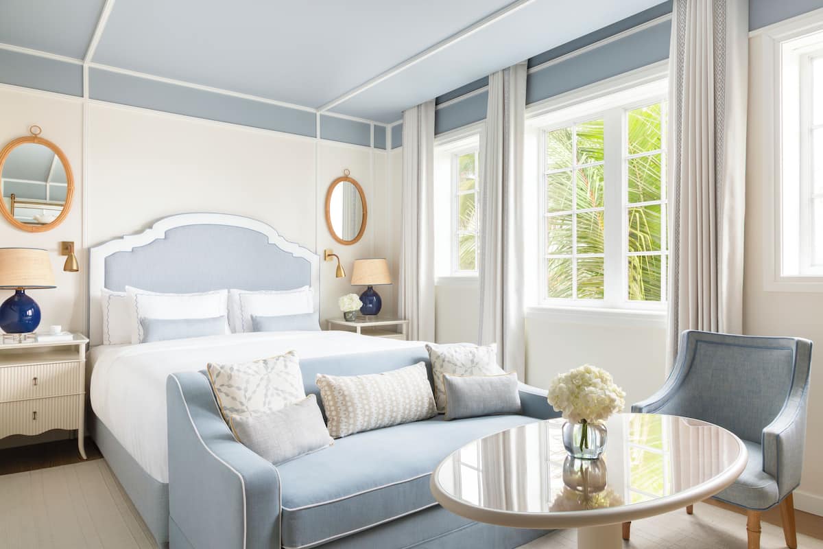 A hotel room with a large bed, a round table with a vase, a chair, and a window with curtains.