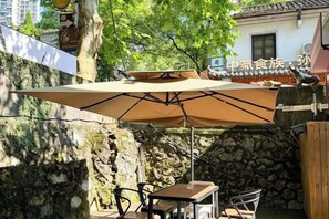 Un patio exterior con mesa y sillas bajo un paraguas grande, rodeado de muros de piedra y follaje.
