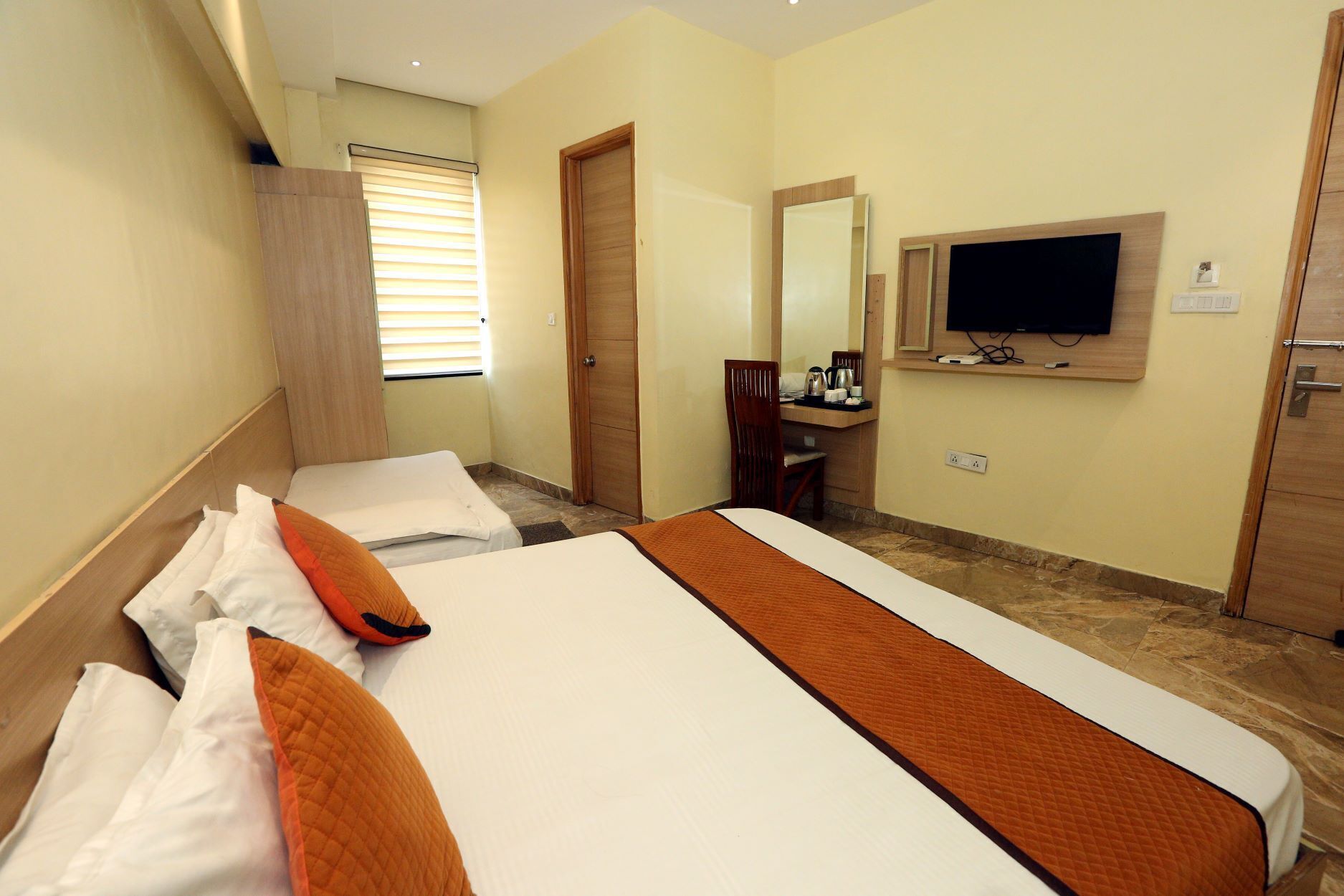 A hotel room with two beds, a desk with a chair, a television mounted on the wall, and a window with blinds.