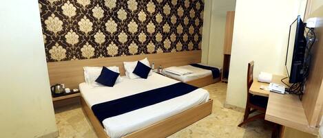 A hotel room with a large bed, a smaller bed, a desk with a chair, a television, and a patterned wallpaper.