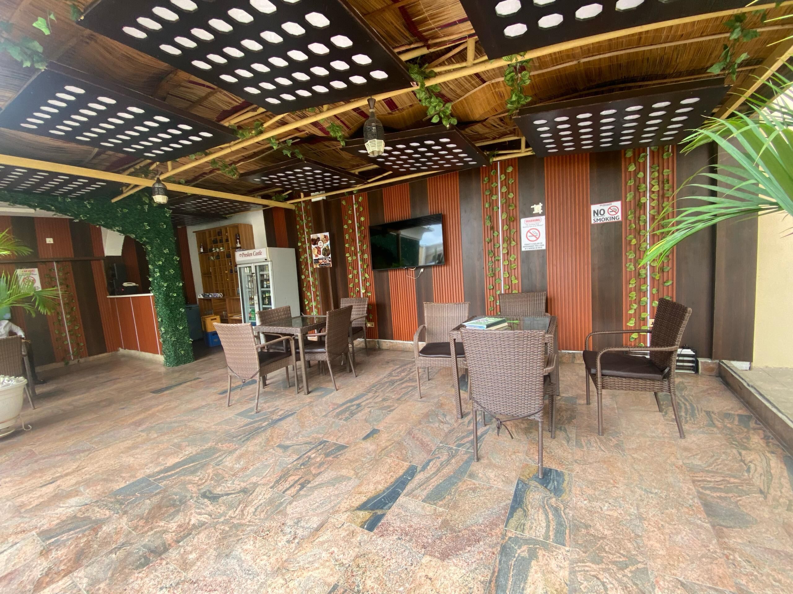 A restaurant with a modern interior, featuring a wooden ceiling, a TV mounted on the wall, and a seating area with wicker chairs and a glass table.